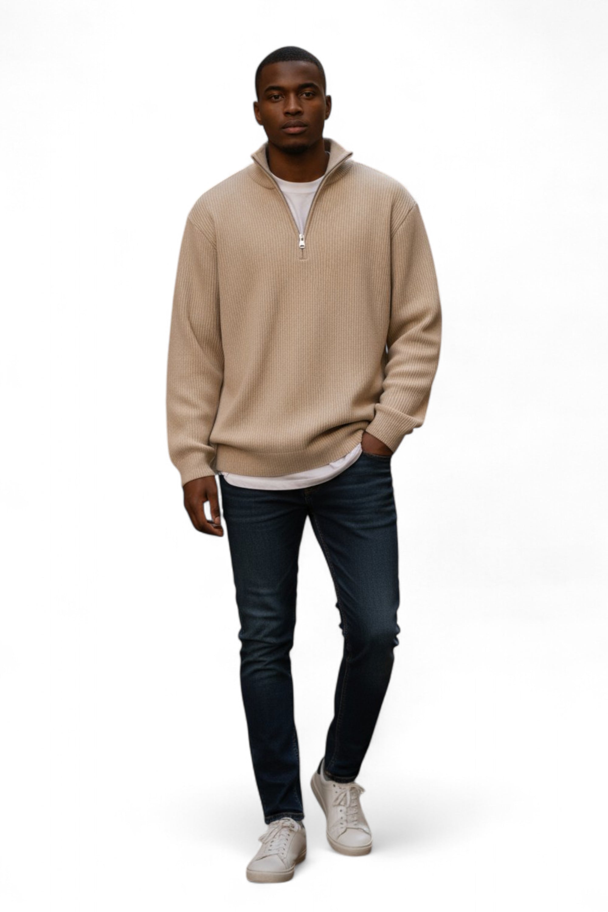 Man wearing a beige sweater, white shirt, dark jeans, and light gray shoes on a white background
